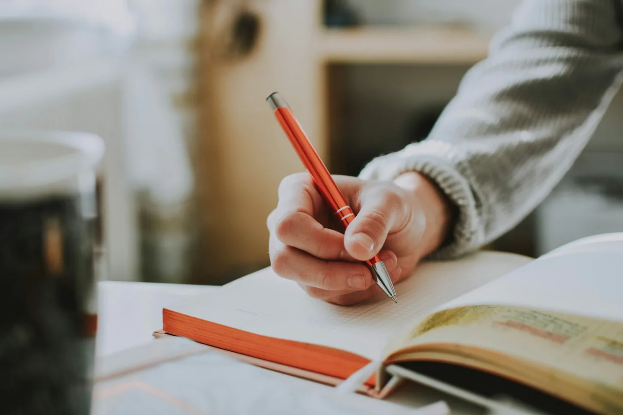 A person writing notes in an open notebook with a red pen.